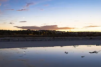 Sunset over coastal village with reflections on shallow water and sandy shoreline.