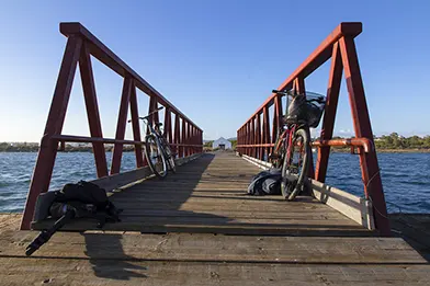 Two bicycles parked on a wooden pier with red railings, overlooking water under a clear blue sky.
