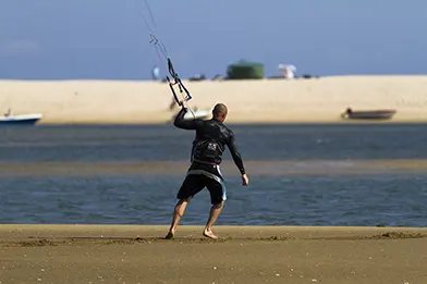 Man kite surfing from sandy shore with kite control bar, shallow water and sand dunes in background.
